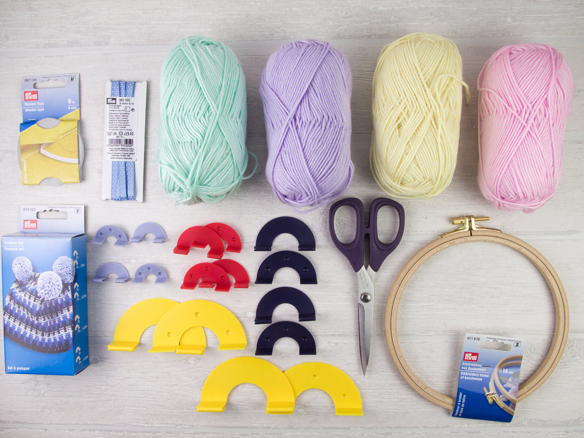 Materialien für ein selbstgemachtes Pompon-Mobile: Bunte Wolle, Stickrahmen aus Holz, Pomponschablonen, Stickgarn und Schere für ein kreatives Bastelprojekt mit Pompons.