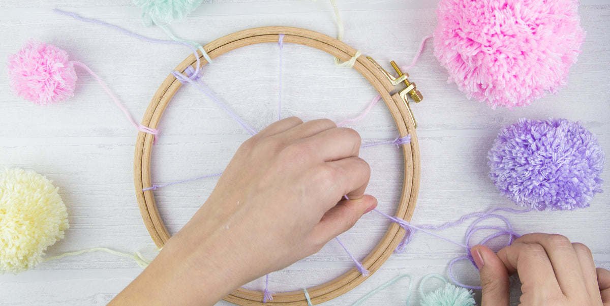 Ein Pompon wird mit Wollfäden an einem Stickrahmen festgeknotet.