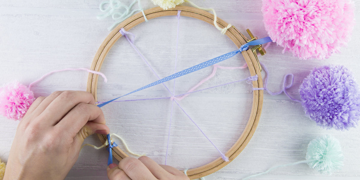 Ein Satinband wird als Aufhängung an einem mit Pompons dekorierten Stickrahmen befestigt.